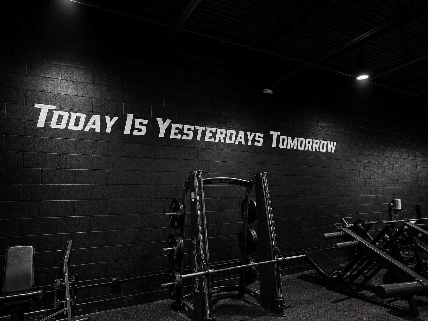 A Smith machine and weight benches set against a black wall painted with the words Today Is Yesterdays Tomorrow.